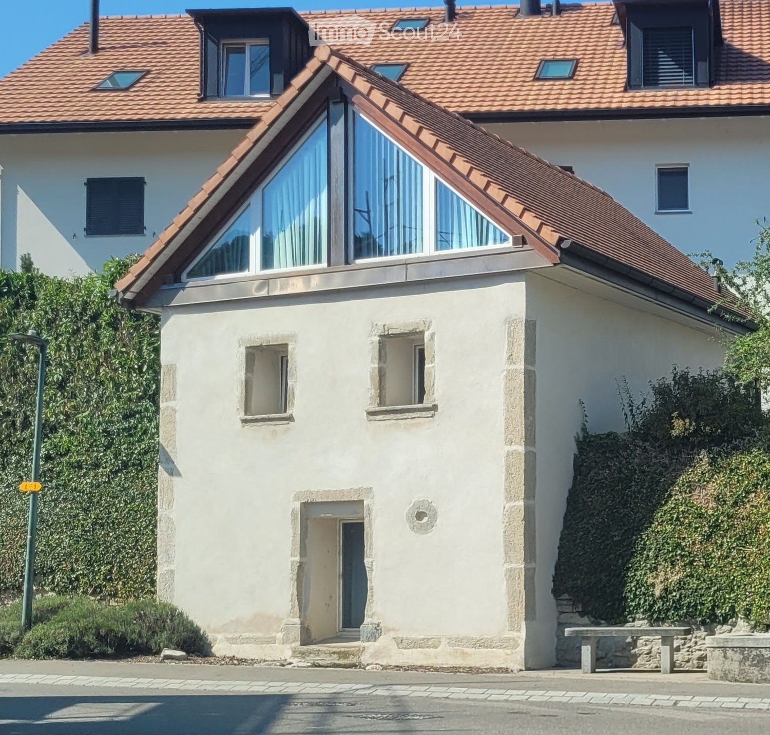Two-story house with a red-tiled roof, white walls, and a modern glass extension on the roof. Features include a front door with steps, small windows, and a stone bench in the garden area.
