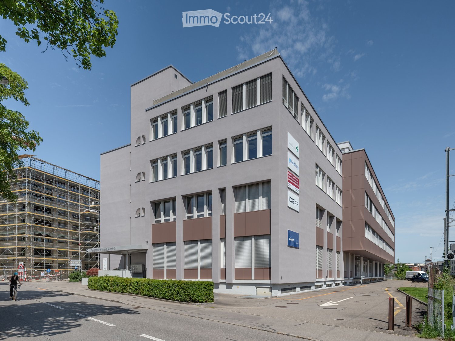 Modern multi-story building with large windows and brown accents, featuring signage for "Arbeitszentrum Liebfeld" and other companies. The building has a clean, contemporary design with a mix of light and dark tones.