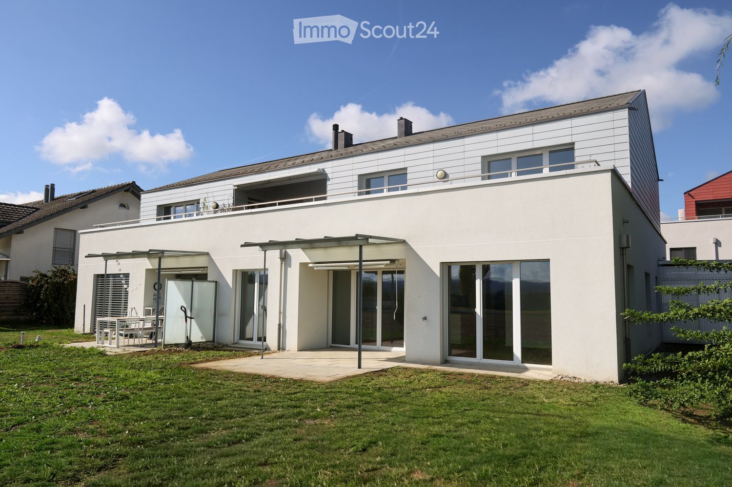 Modern two-story house with white exterior, large windows, and a balcony on the upper level. The house features a covered patio area with outdoor furniture and a well-maintained lawn.