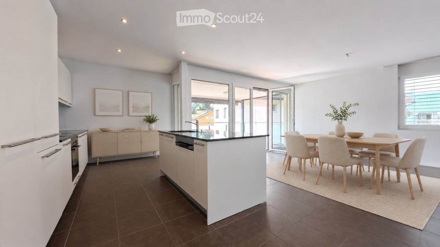 Modern kitchen and dining area with white cabinets, a central island, and a dining table with chairs. Large windows and sliding doors lead to a balcony.