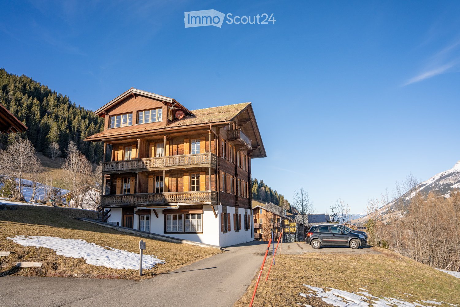 A two-story wooden house with a balcony, surrounded by a grassy area and snow patches, with mountains in the background.