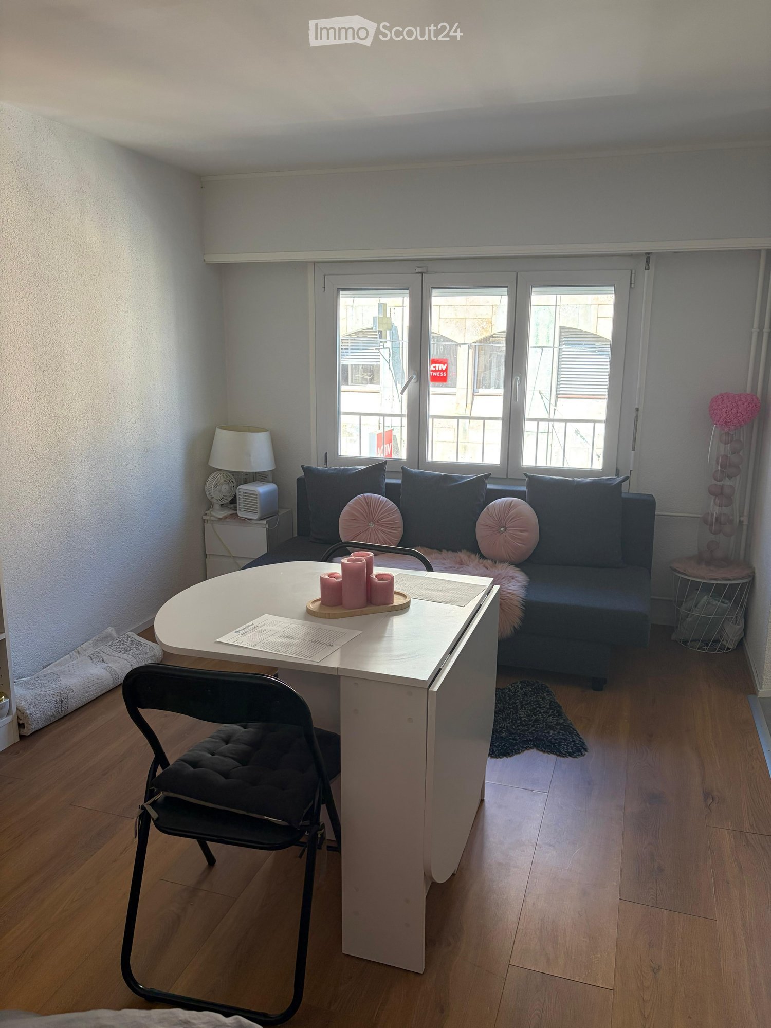A small, well-lit room with a sofa, a round table, a chair, a lamp, and a fan. The room features wooden flooring and large windows with blinds.