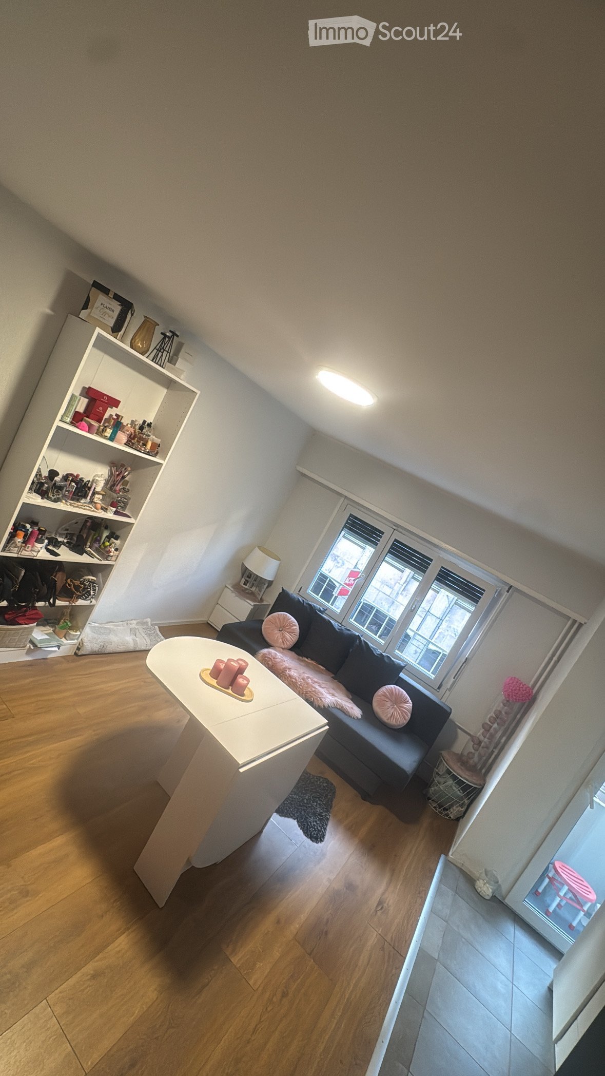 Attic living room with wooden flooring, a dark blue sofa with pink cushions, a white coffee table with candles, a shelf with various items, and a small side table with a lamp.