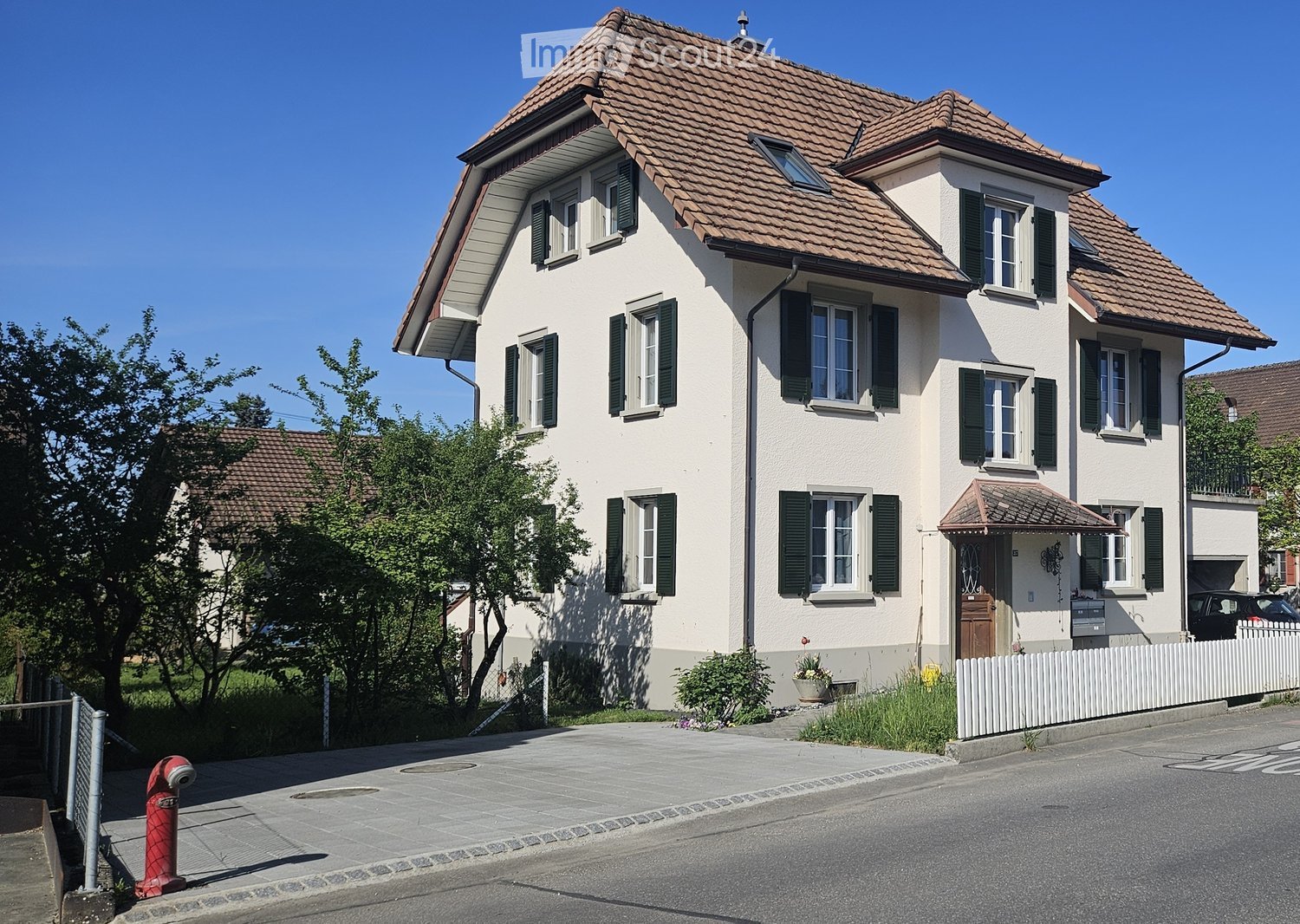 House. Two-story house with a tiled roof, white exterior walls, green shutters, and a small garden area with a white picket fence.