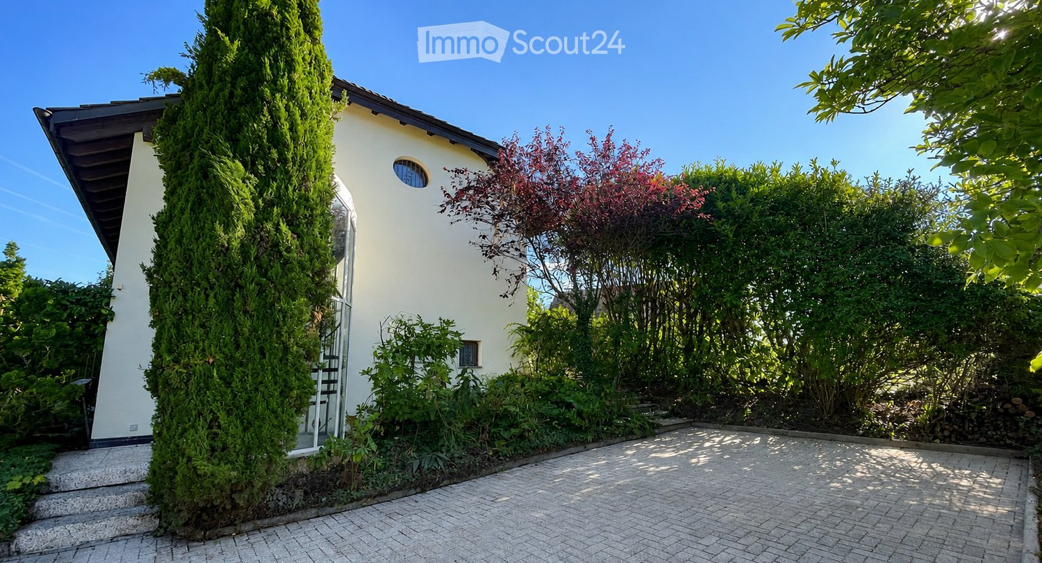 A modern house with a well-maintained garden, featuring a tall green conifer, various shrubs, and a paved pathway leading to the entrance.