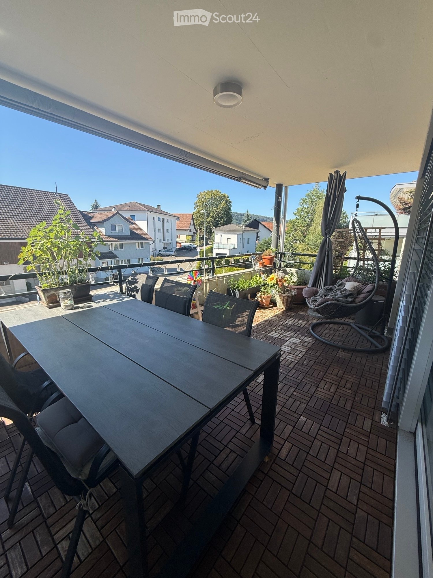 Balcony with a wooden table, chairs, a hanging wicker chair, potted plants, and an umbrella. The view shows a residential area with houses and trees.
