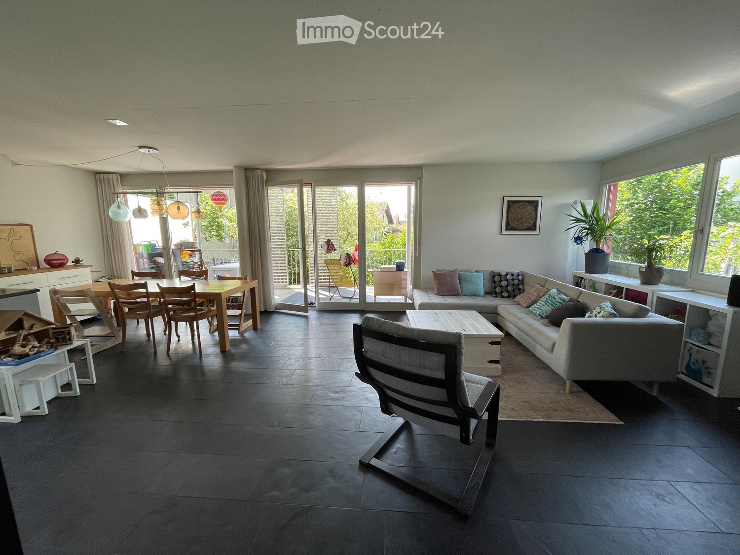 Open-plan living and dining area with dark tiled flooring, large windows, and a balcony access. Features a grey sectional sofa, coffee table, dining table with chairs, pendant lights, and shelves with decorative items.