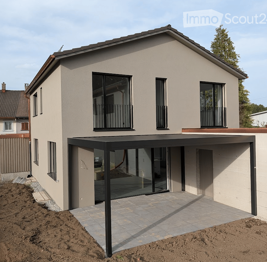 Modern two-story house with beige facade, large windows, and balconies. Features a covered patio area with glass walls and a tiled floor.
