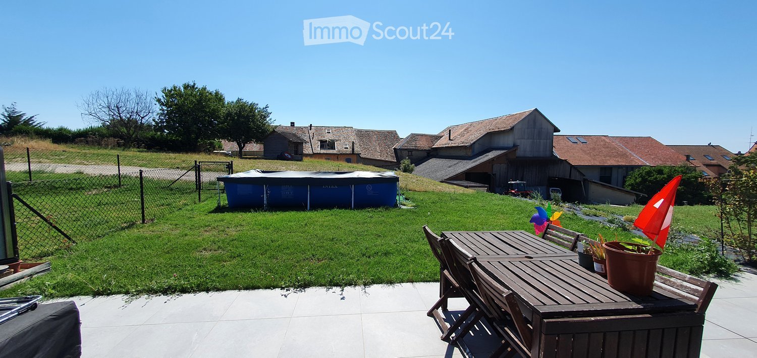 A well-maintained garden with a wooden patio table and chairs, potted plants, and a colorful pinwheel. The background features a grassy lawn, a fenced area, and several houses with tiled roofs.