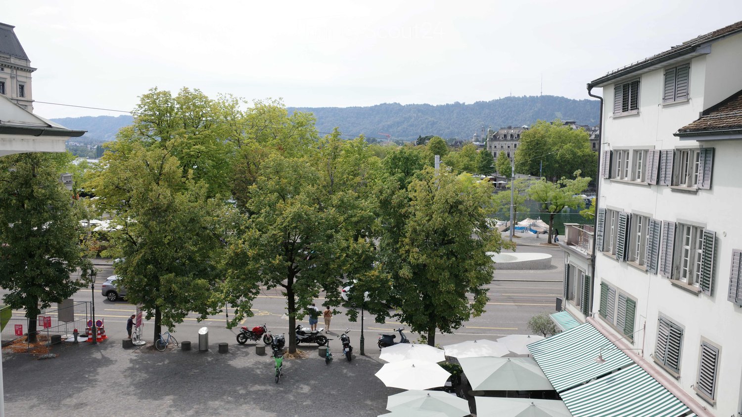 Balcony view of a city square with trees, parked motorcycles, bicycles, and outdoor seating umbrellas.