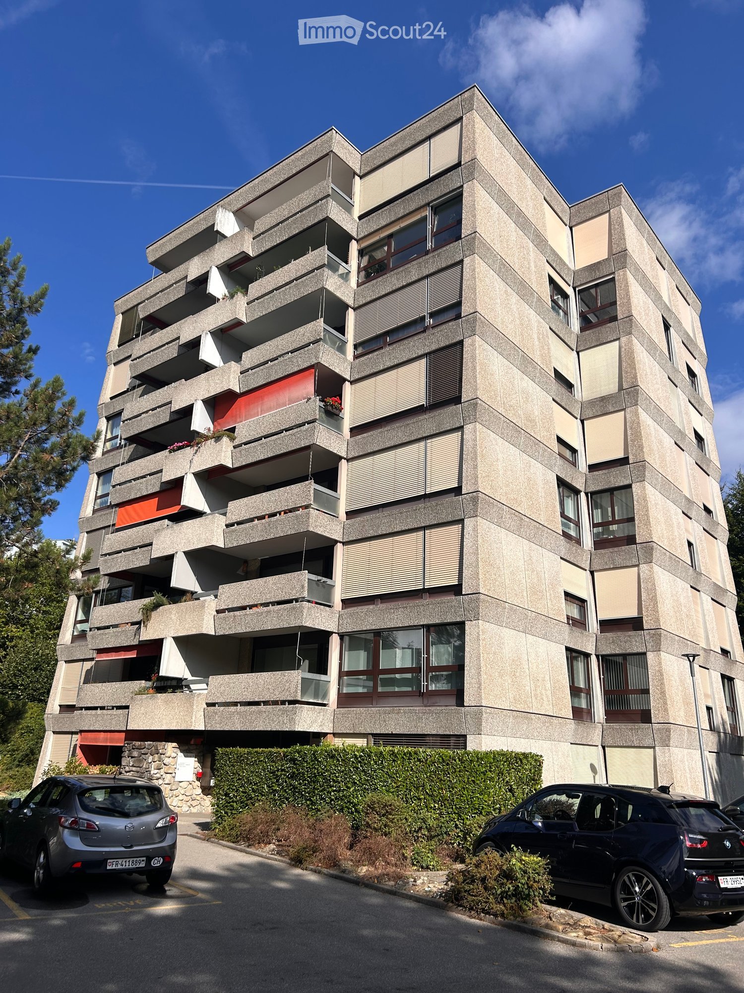 Modern multi-story apartment building with concrete facade, balconies, and large windows. Two cars are parked in front.