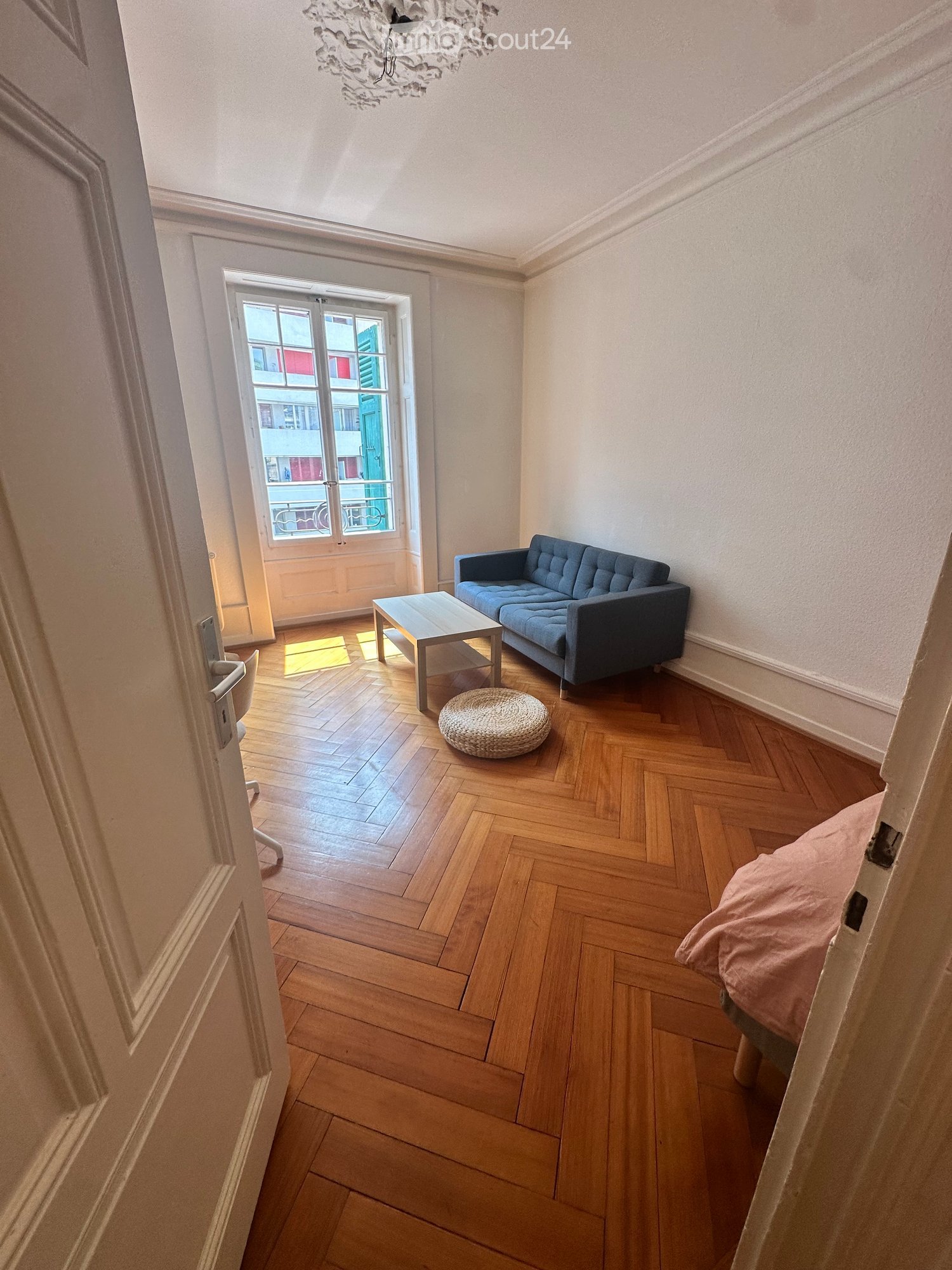 A small, well-lit living space with wooden herringbone flooring, a blue sofa, a coffee table, a round woven stool, and a bed with a pink blanket visible through an open doorway.