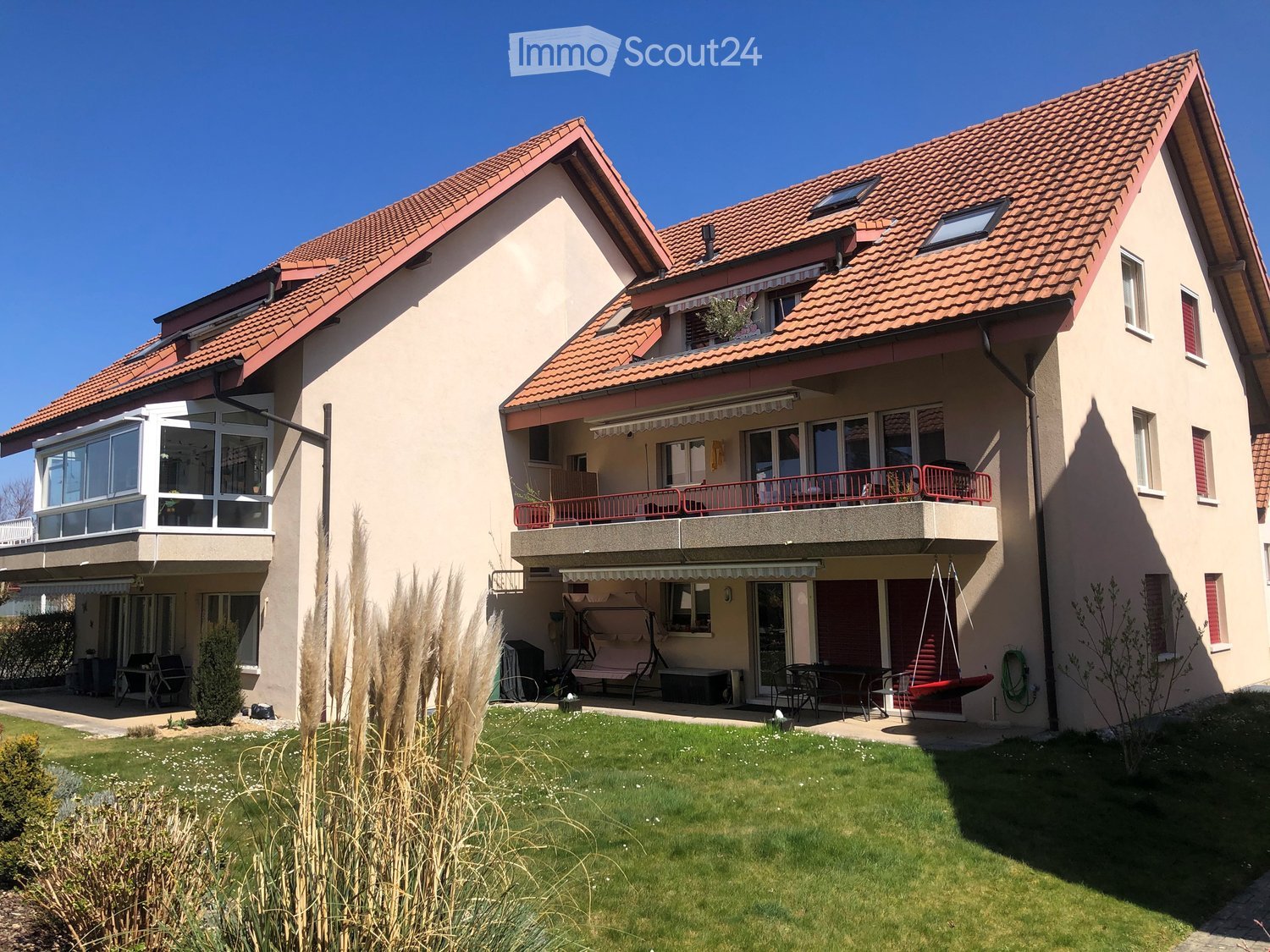Two-story house with red-tiled roof, beige facade, large windows, balcony, and well-maintained garden.