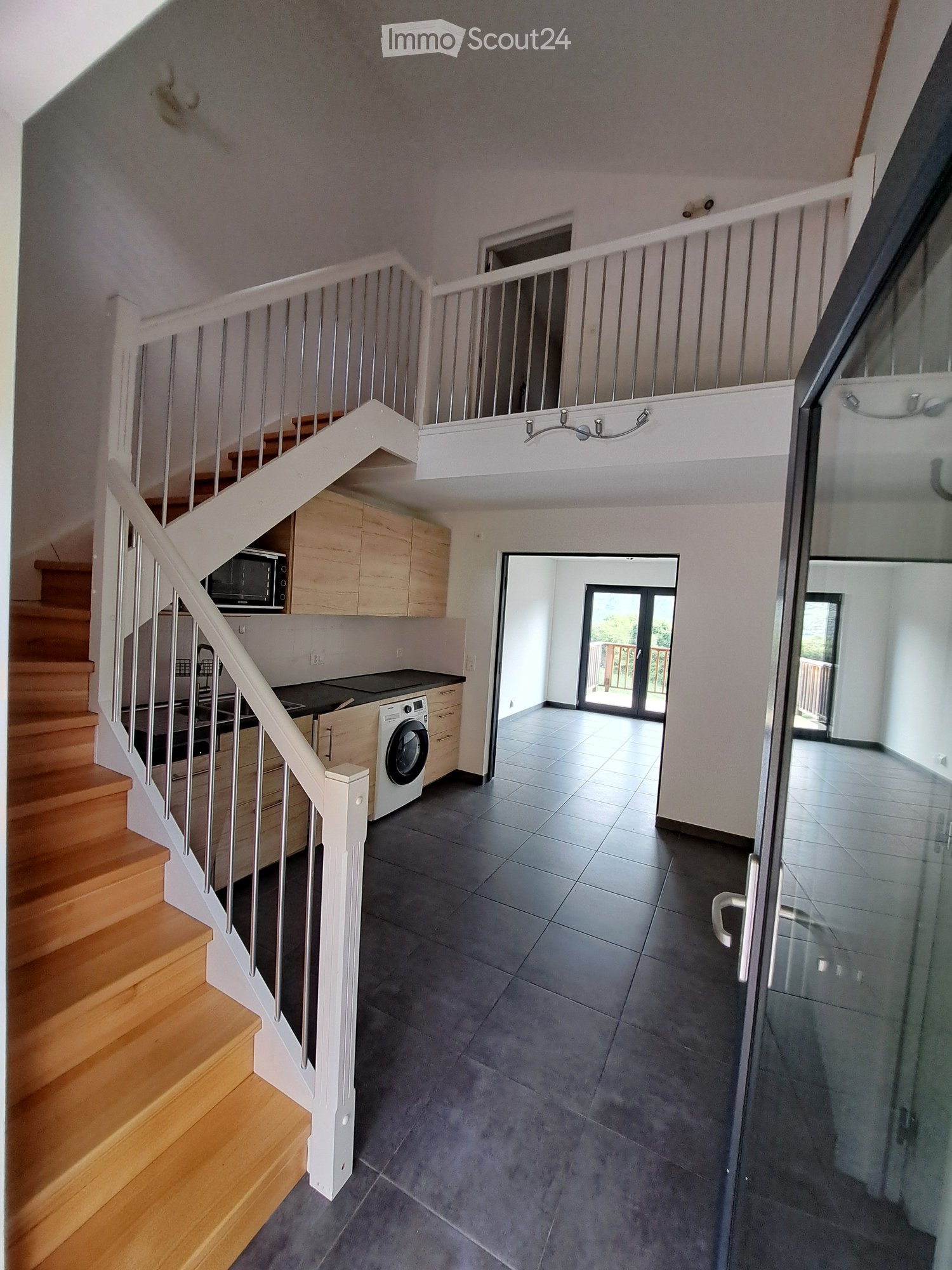 Modern hallway with wooden staircase, white railing, grey tiled floor, and open kitchen area visible with cabinets, microwave, and washing machine.