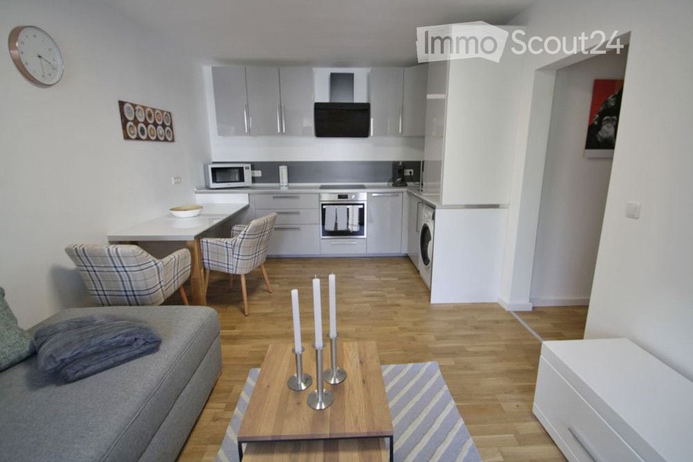 Modern open-plan living and dining area with a grey sofa, coffee table, dining table with chairs, and a fully equipped kitchen featuring a microwave, oven, and washing machine.
