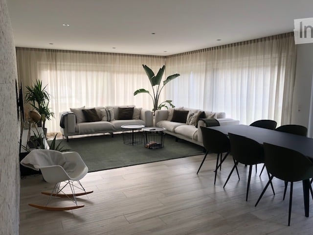 Modern living room with a grey sectional sofa, coffee tables, a white rocking chair, and a dining table with chairs. Large windows with sheer curtains and indoor plants add to the decor.