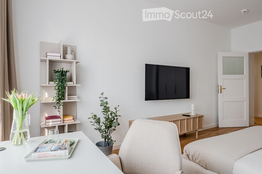 A modern, well-lit room with a bed, chair, small table, shelf with books and plants, and a television mounted on the wall. The room features light-colored walls and wooden flooring.