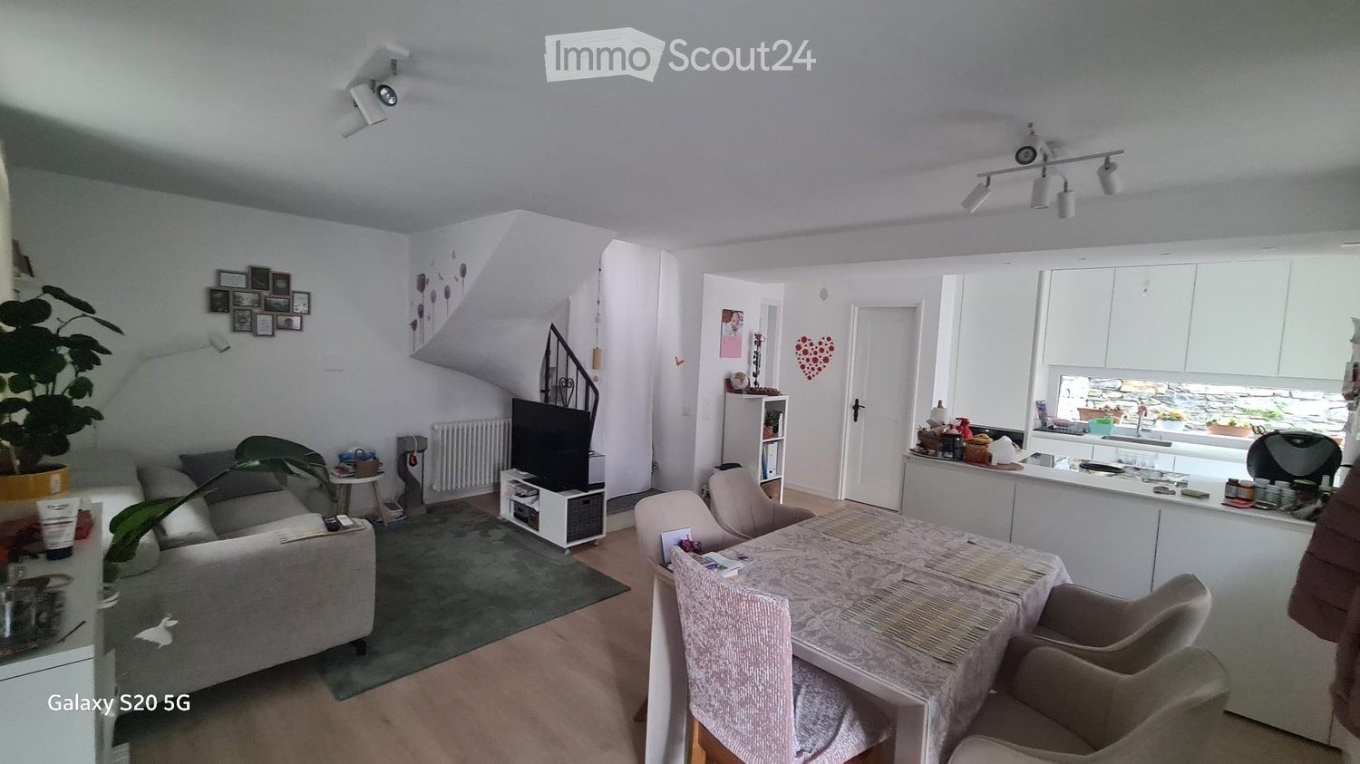 A modern, well-lit living and dining area with a grey sofa, a dining table with chairs, a television, and a kitchen area with cabinets and a sink.