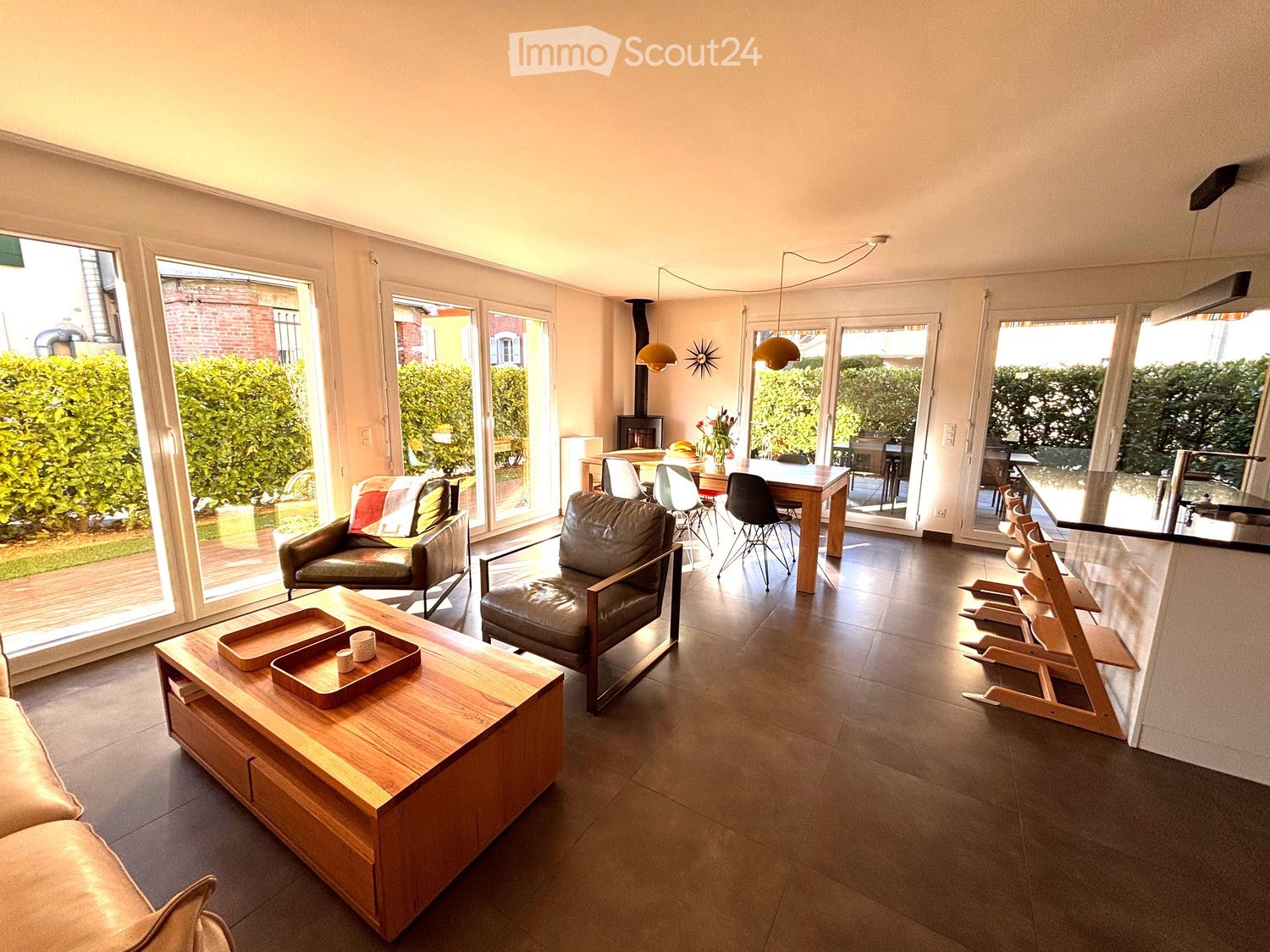 Bright living room with large windows and sliding doors leading to a patio, featuring a sofa, coffee table, armchairs, dining area, and fireplace.