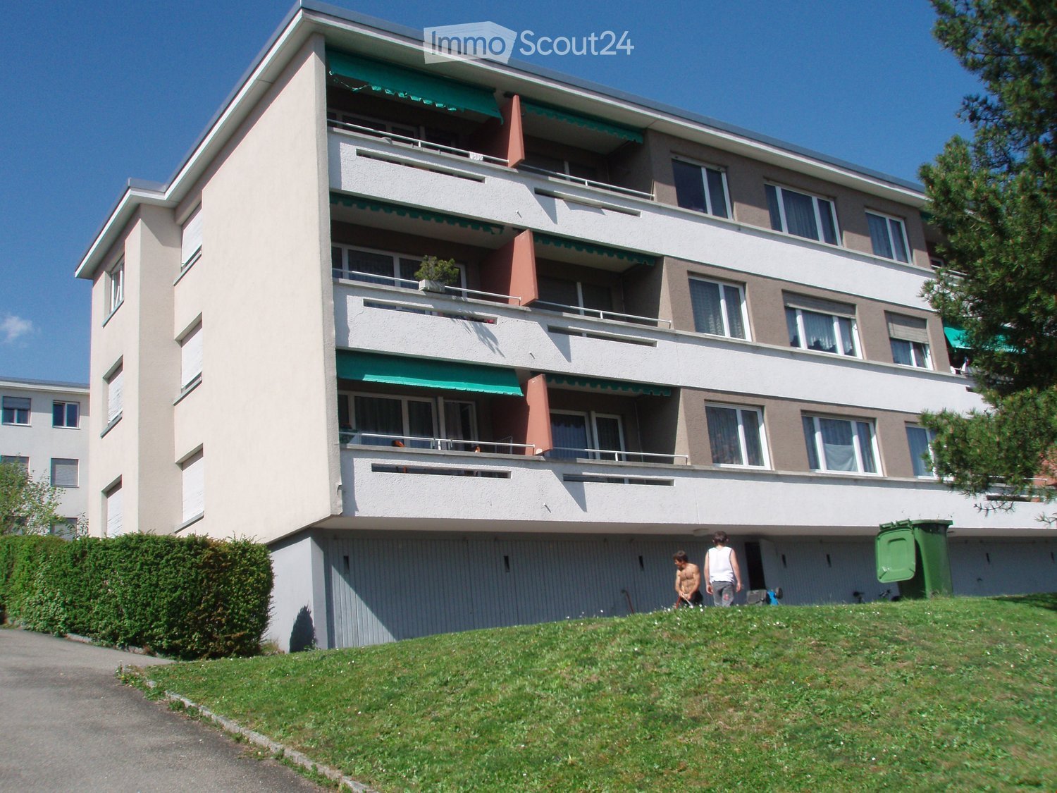 Multi-story residential building with balconies, green awnings, and a raised ground floor. Two people are visible near the entrance.