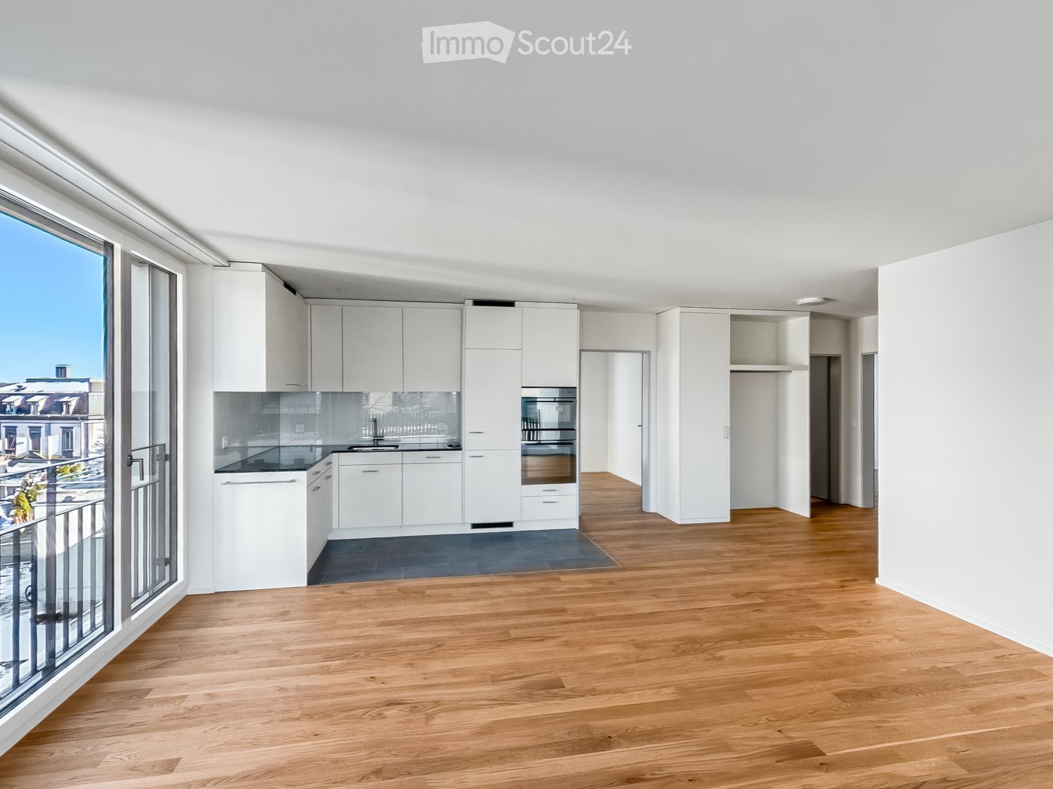 Open-plan unfurnished room with wooden flooring, white cabinetry, and large windows leading to a balcony.
