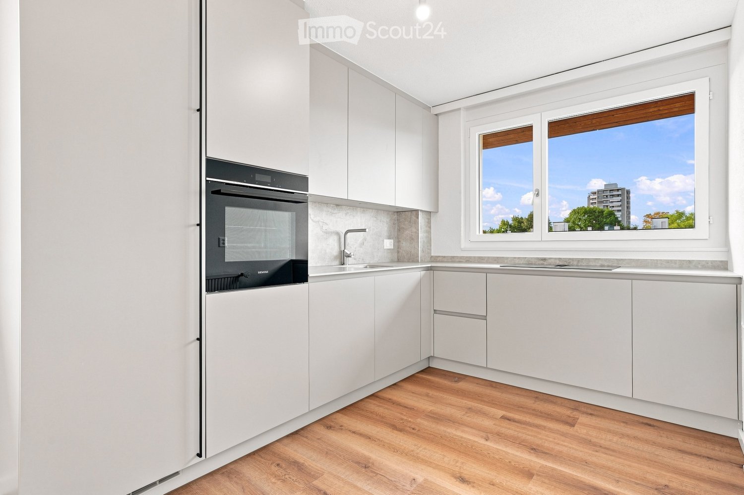 Modern kitchen with white cabinets, wooden flooring, and a large window overlooking a cityscape.