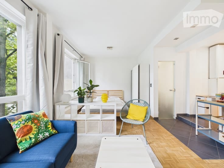 Bright, airy room with large windows offering a view of greenery. Features a blue sofa, a white table, a bed, a chair with a yellow pillow, and several potted plants.