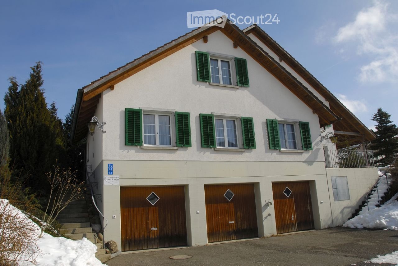 Two-story house with white walls, green shutters, wooden garage doors, and a snow-covered driveway.