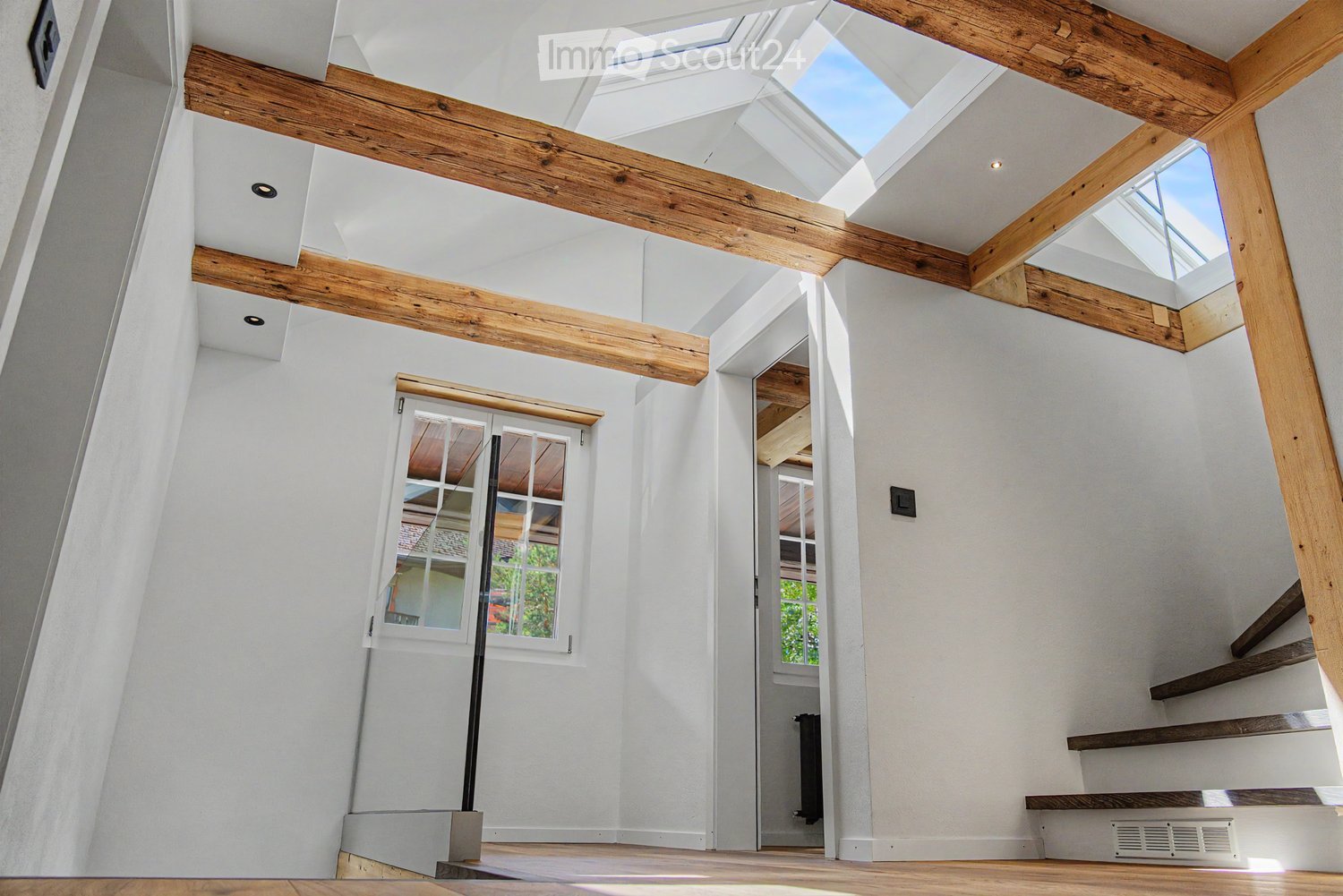 Bright, airy hallway with exposed wooden beams, skylights, and a staircase leading upwards. White walls and wooden flooring.