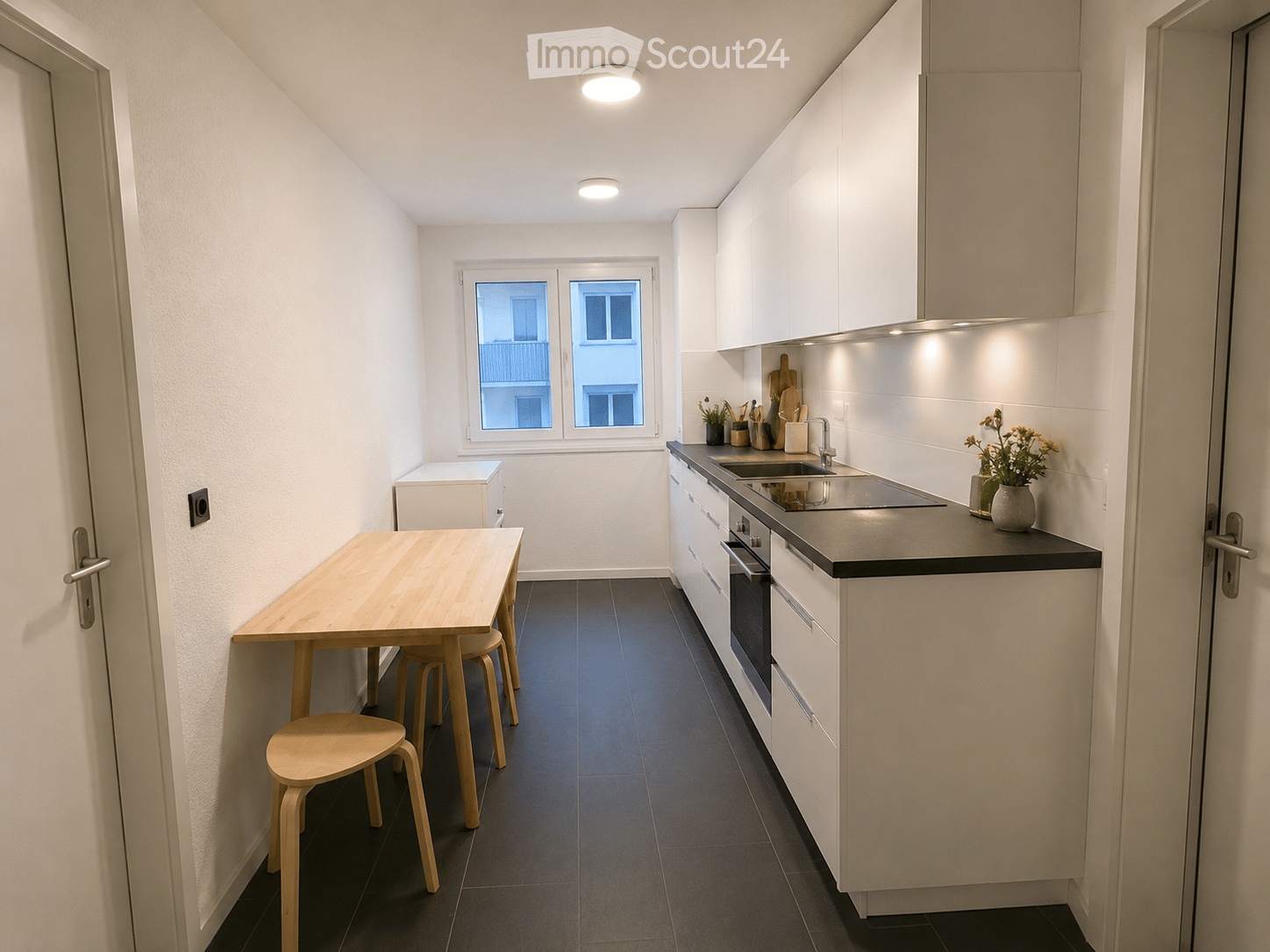 Modern kitchen with white cabinets, dark countertops, a wooden dining table with four stools, and a window overlooking a cityscape.