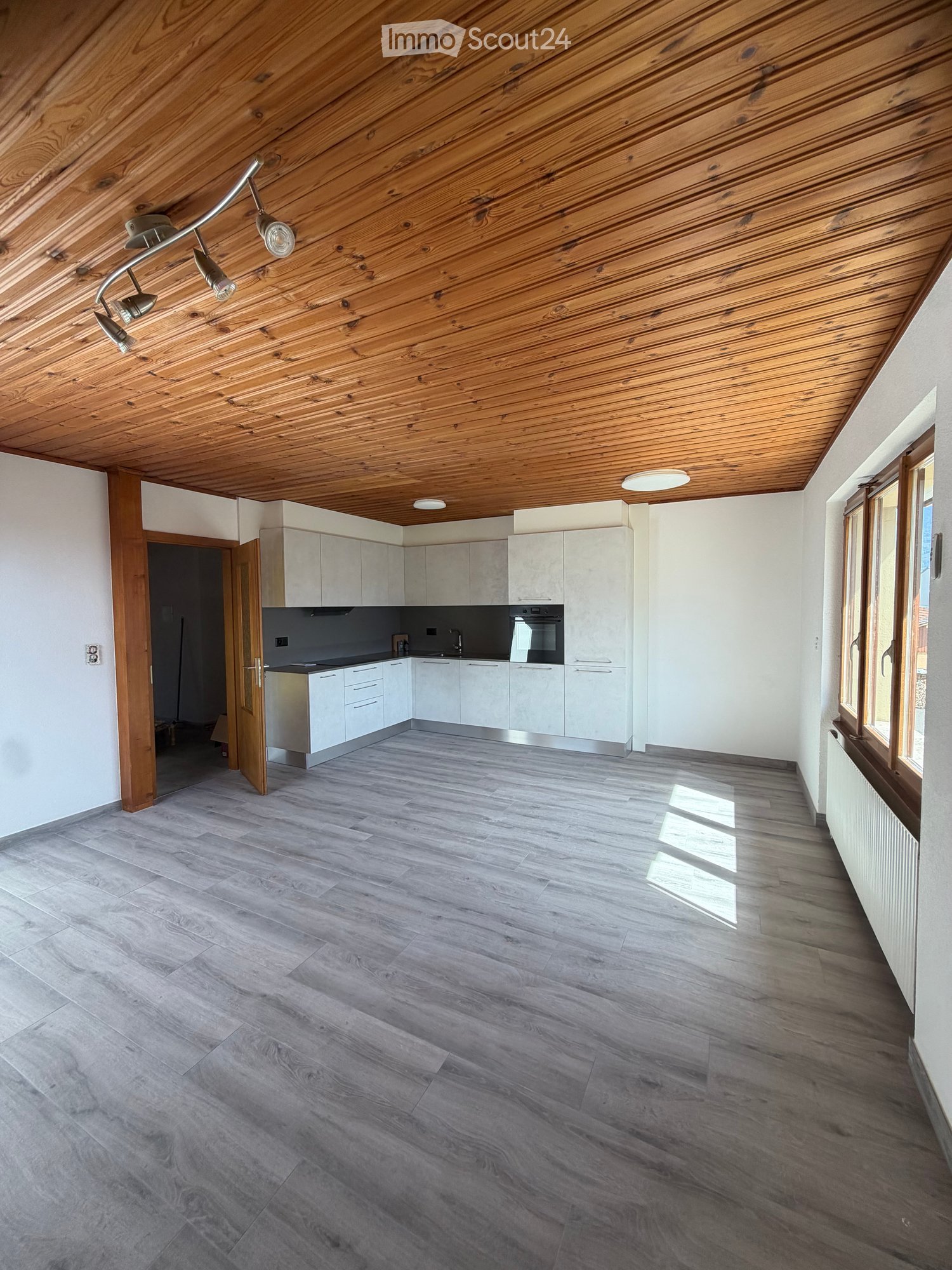 Empty room with wooden ceiling, grey wooden flooring, white walls, and large windows. Kitchen area visible with white cabinets and stainless steel appliances.