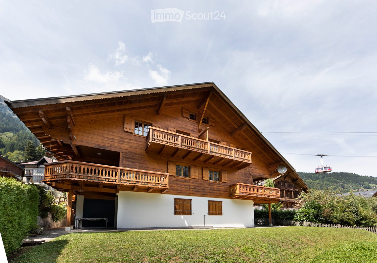A large, multi-story wooden house with balconies and a spacious lawn in front, set in a scenic mountainous area.