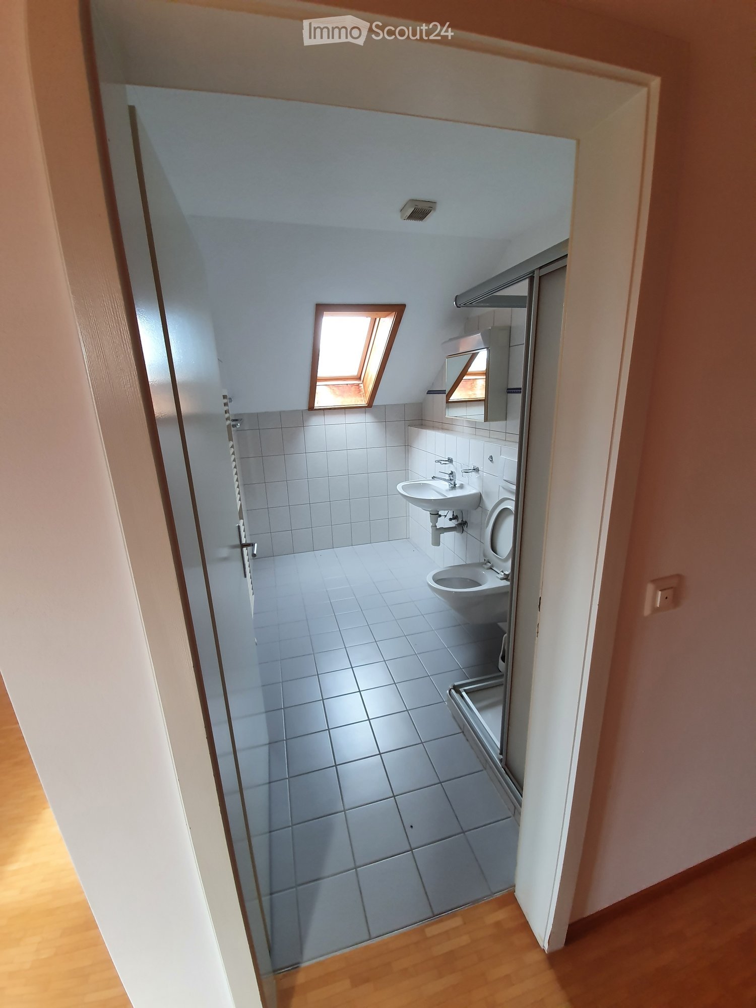 Small, clean bathroom with white tiled walls and floor, a sink, toilet, mirror, and a small shower area with a glass door. Natural light comes from a skylight window above the toilet.