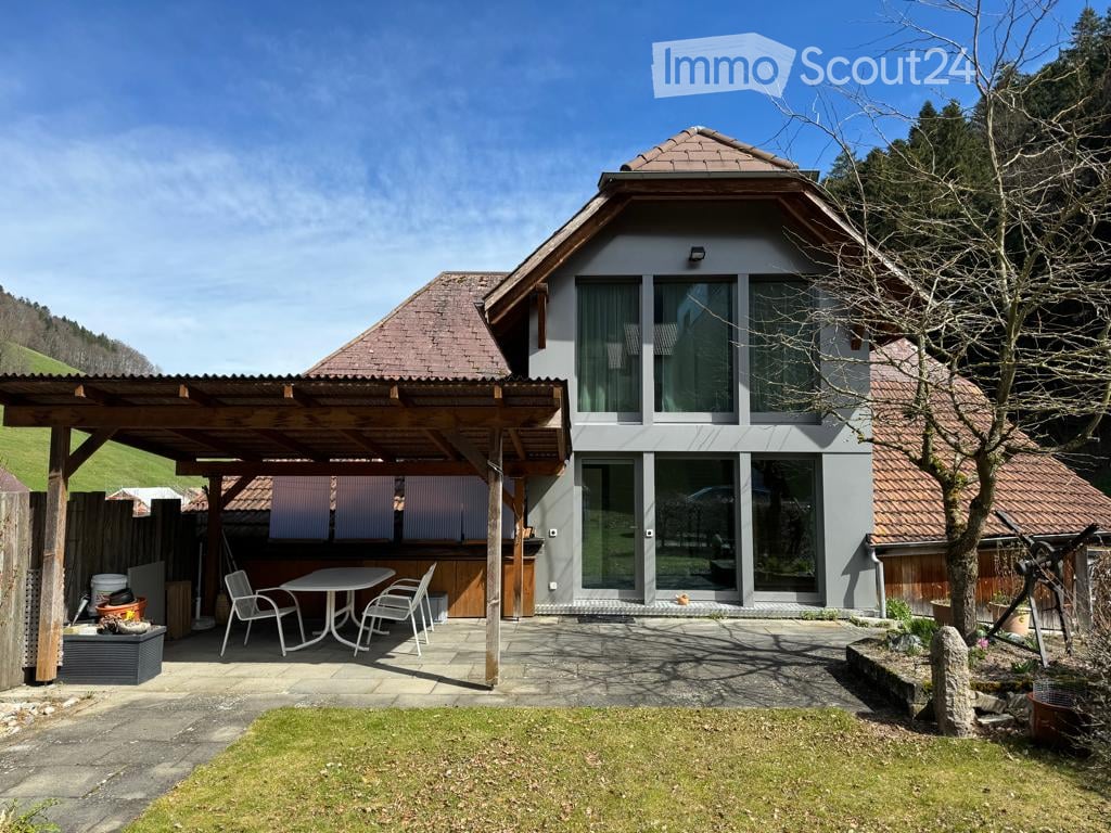 A modern two-story house with a wooden pergola and outdoor seating area, surrounded by a well-maintained garden and scenic hillside views.