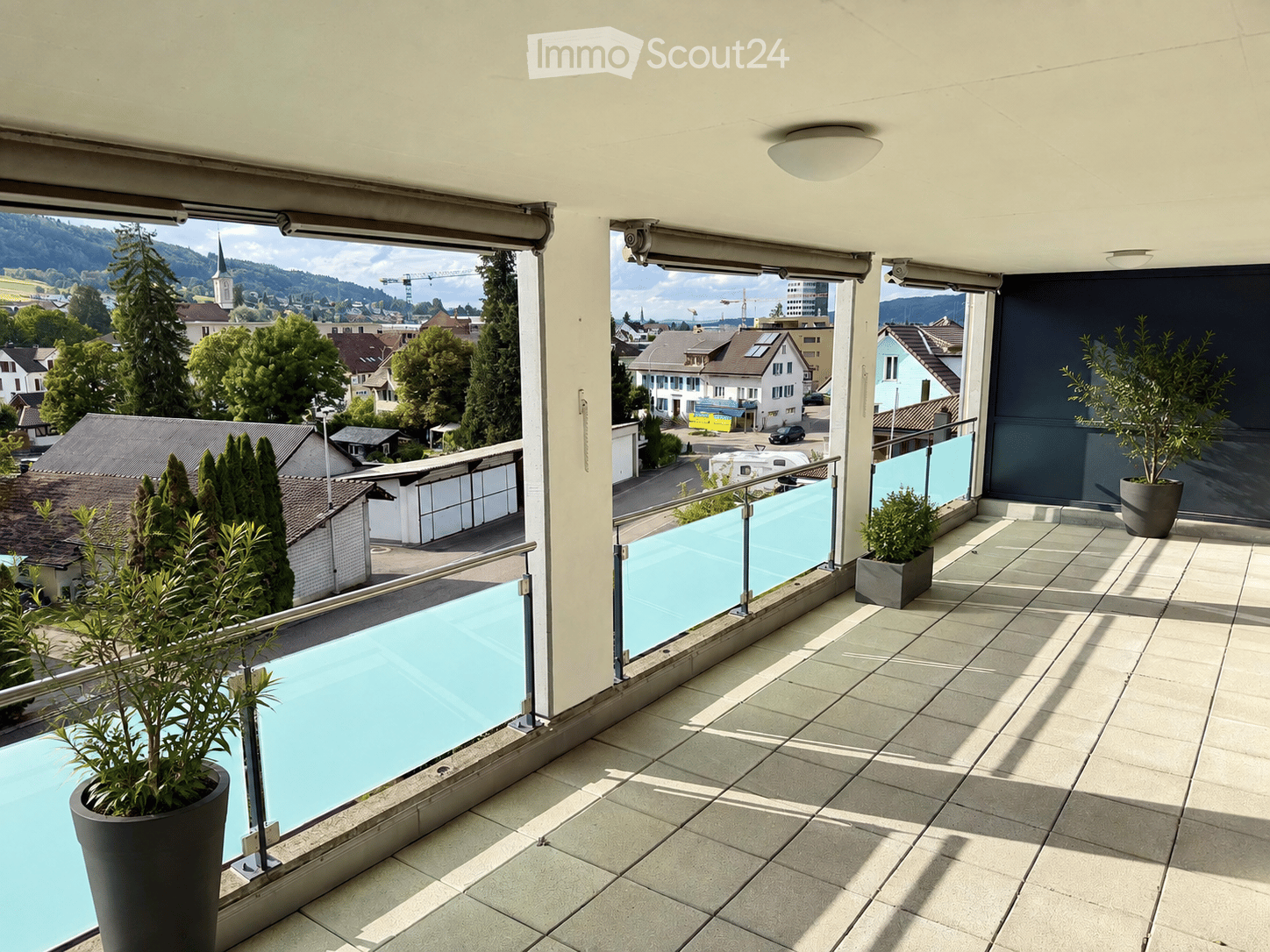 Balcony with tiled floor, glass railing, and scenic city view, featuring potted plants.
