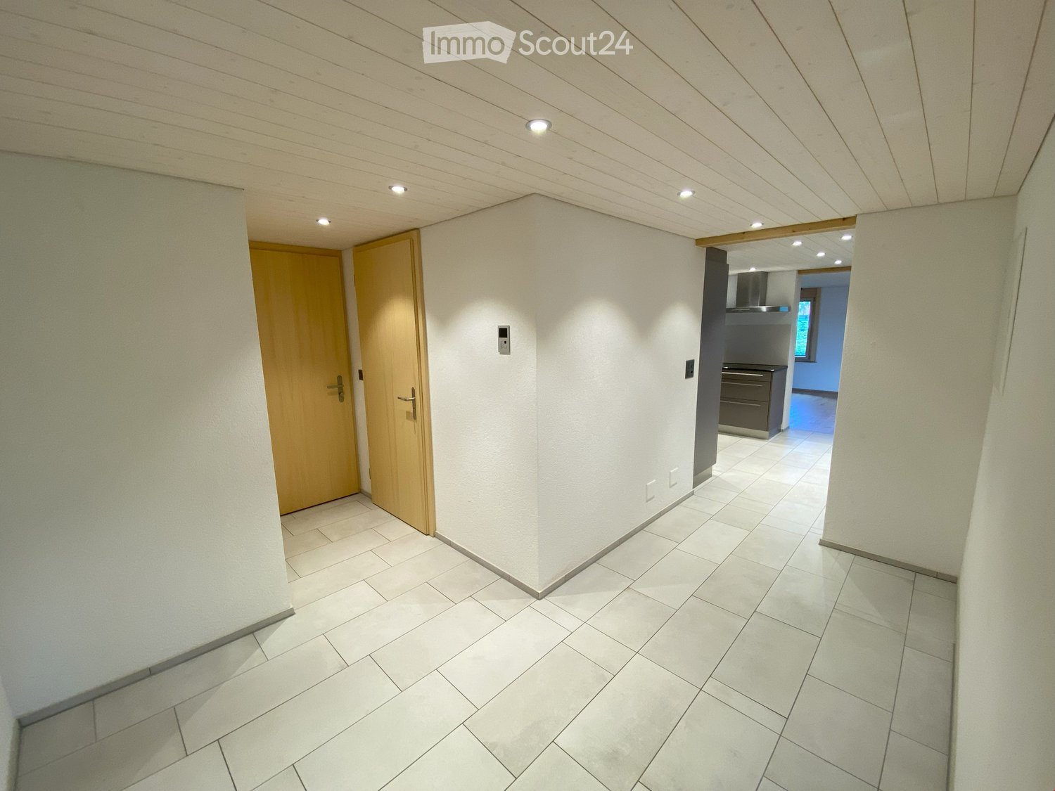 Clean hallway with tiled flooring, white walls, wooden doors, and recessed lighting. A small kitchen area is visible at the end.