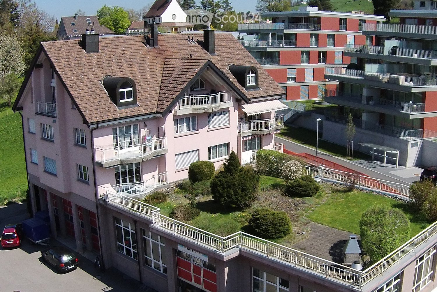 Aerial view of a two-story residential building with a pink facade, multiple balconies, and a well-maintained garden area. The building features a garage and parking area, with neighboring structures visible in the background.