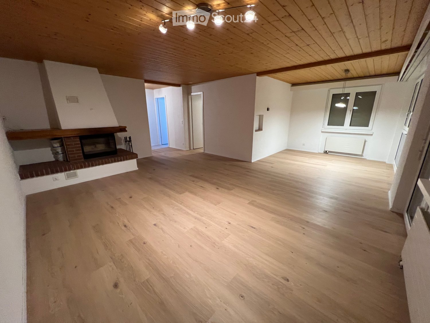 Empty room with wooden flooring, white walls, and a fireplace on the left side. The room has exposed wooden beams on the ceiling and a large window on the right side.