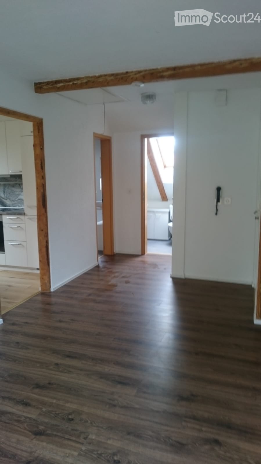 Empty hallway with wooden flooring, white walls, and exposed wooden beams on the ceiling. The hallway leads to other rooms, including a kitchen and a staircase area.
