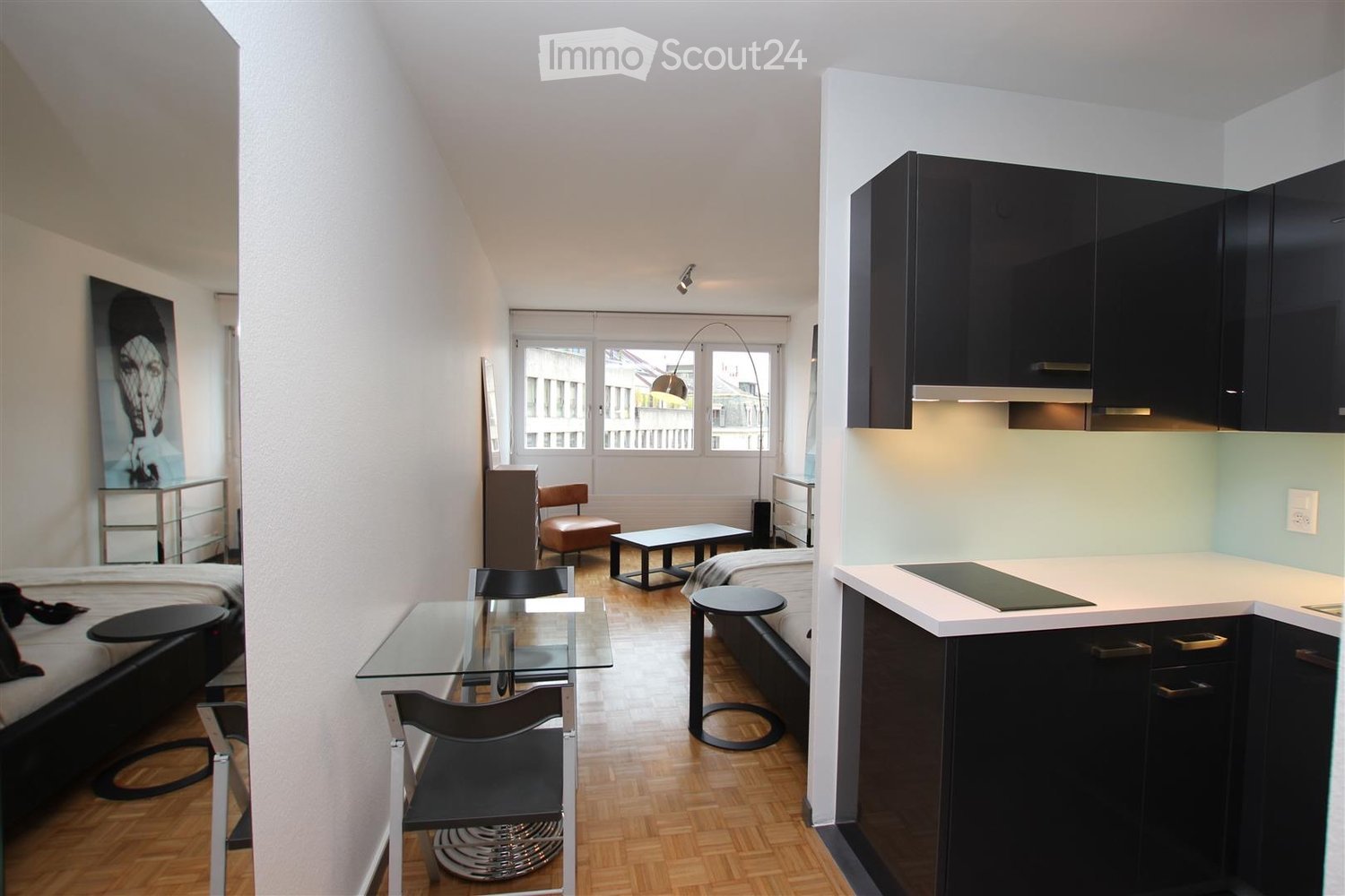 Modern, compact studio apartment with a bed, glass dining table, chairs, a small sofa, and a kitchen area with cabinets and a counter.