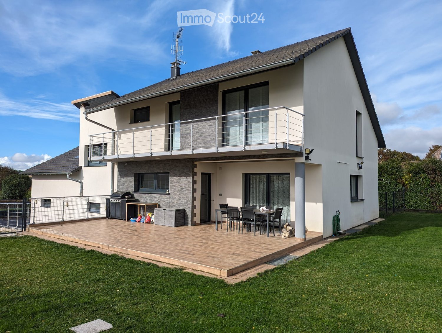 Two-story house with a modern design, featuring a raised ground floor, a balcony, and a well-maintained garden with a patio area and outdoor furniture.