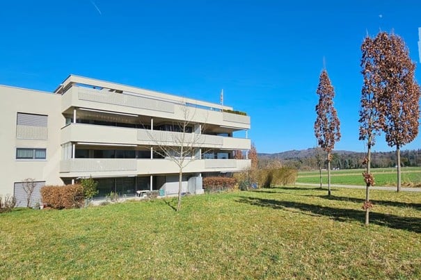 Modern multi-story apartment building with balconies, surrounded by a grassy lawn and trees under a clear blue sky.