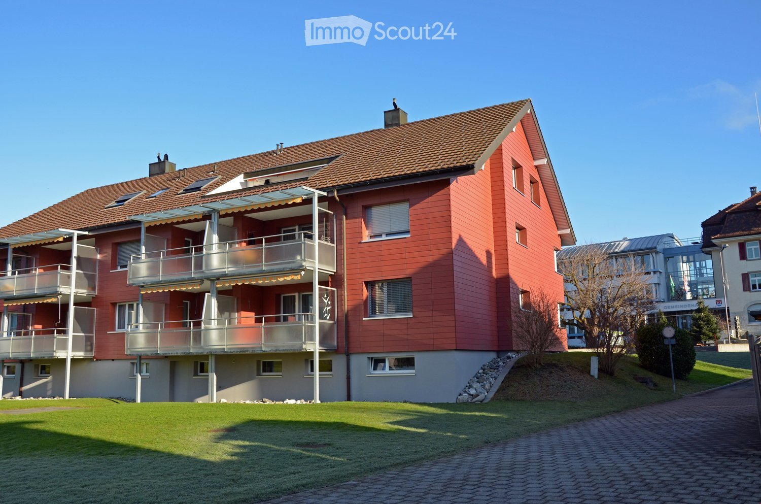 red building with several balconies, gray walls, brown roof, brick ground, garden in front
