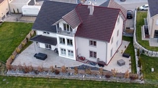 A two-story house with a red-tiled roof, white exterior, and a covered patio area. The house features a garage and a parking area with several cars visible.