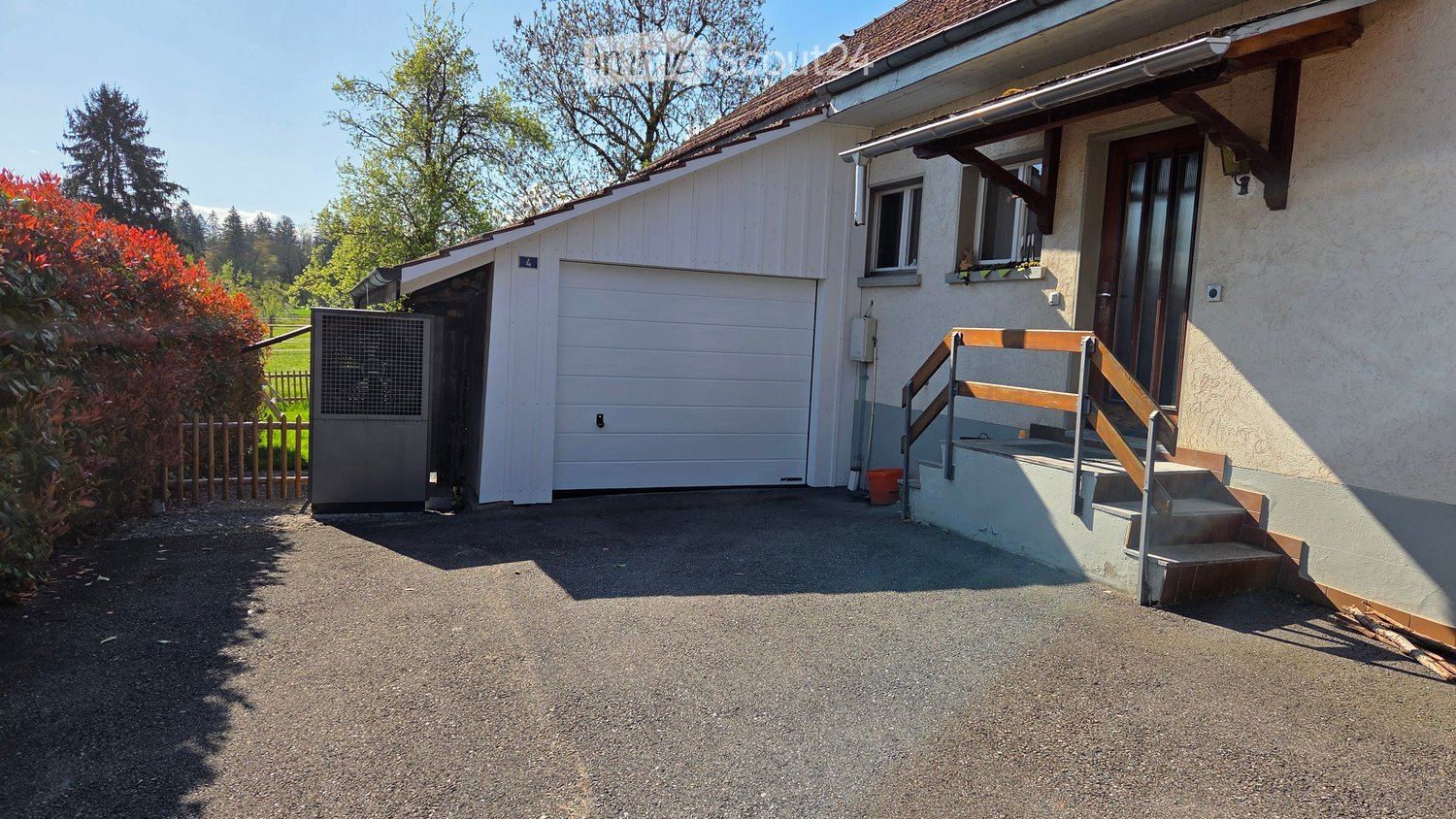 Two-story house with a garage, raised ground floor, and a staircase leading to the entrance. The house has a white exterior with a brown roof and is surrounded by greenery.