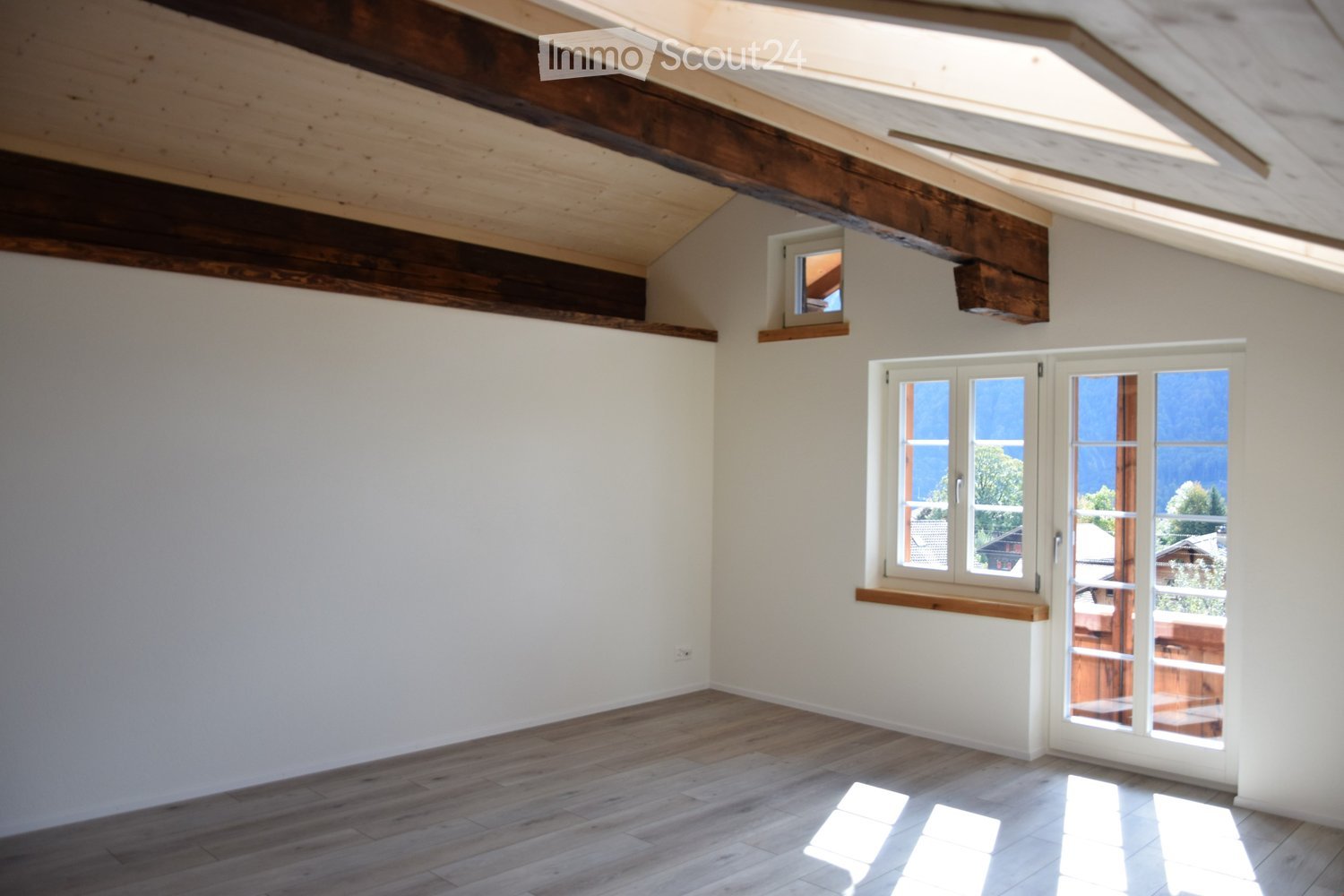 Empty room with exposed wooden beams on the ceiling, light-colored walls, wooden flooring, and large windows offering a scenic view.