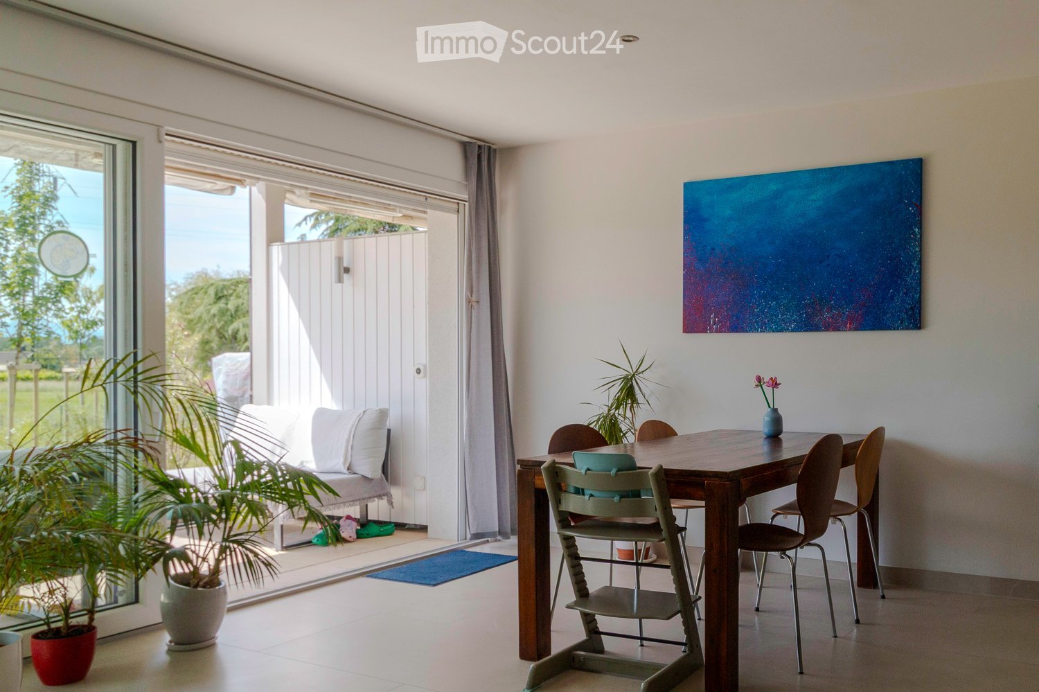 Bright living room with a sofa, dining table, chairs, and a large painting on the wall. Large windows and sliding doors lead to a patio area with additional seating.