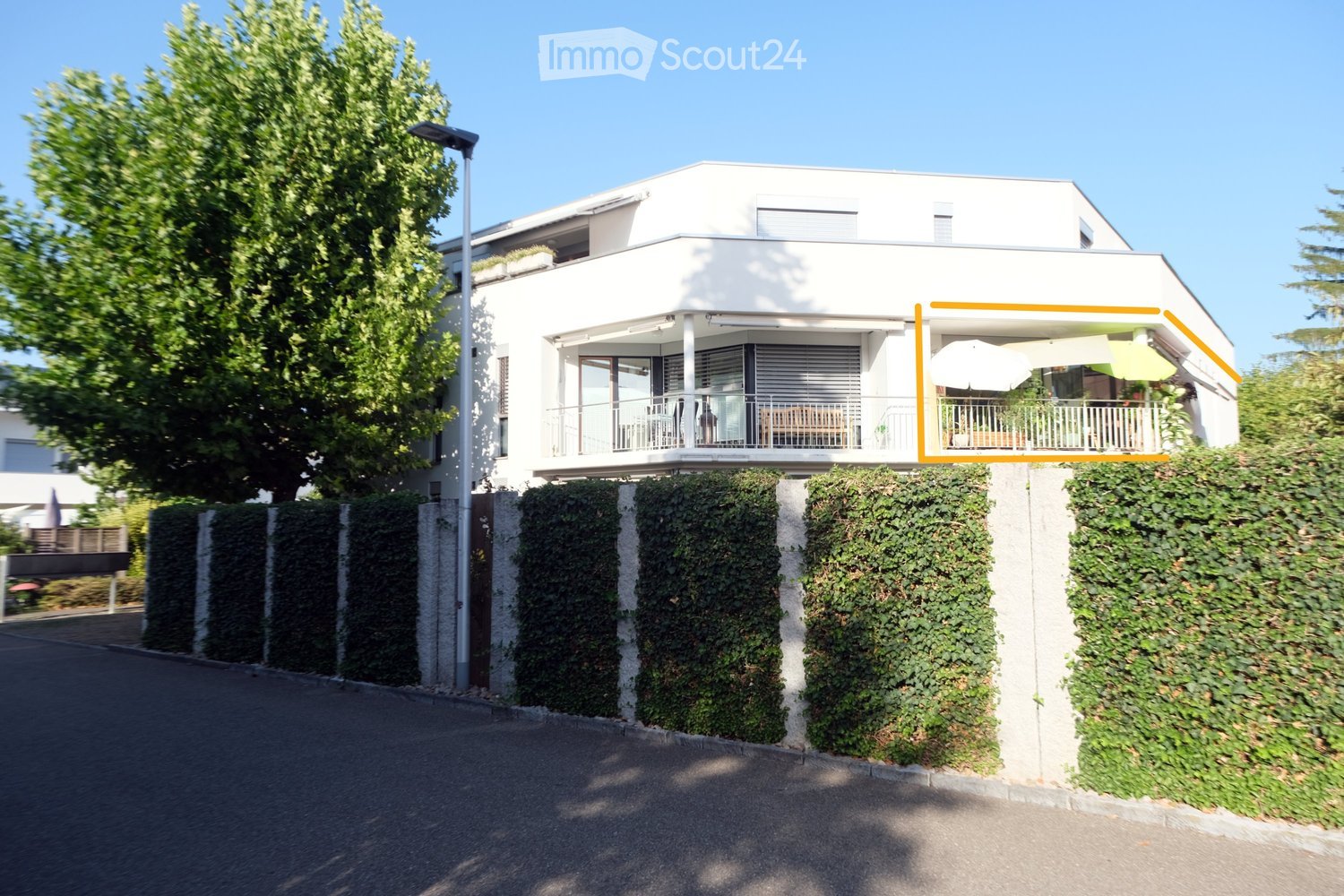 Modern two-story white house with a raised ground floor, featuring a balcony with outdoor furniture and an umbrella, surrounded by greenery and a hedge.