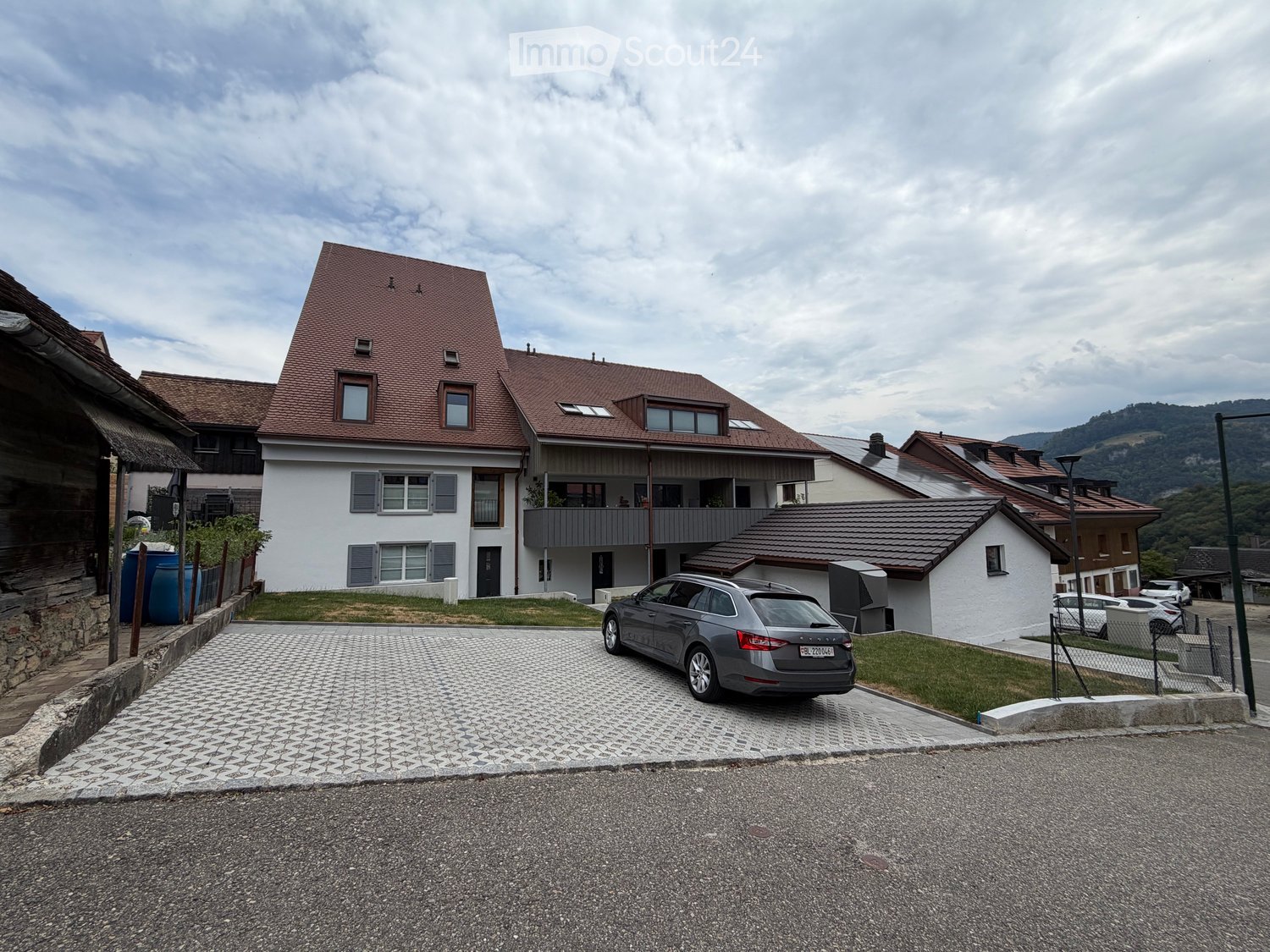 Two-story house with a red-tiled roof, white walls, multiple windows, and a balcony on the upper floor. A grey car is parked in the driveway.
