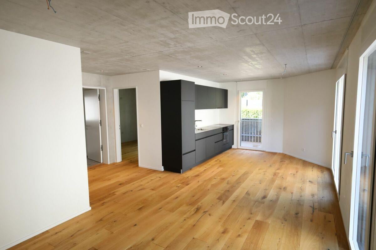 Bright, open-plan living space with wooden flooring, white walls, and a modern kitchen area featuring grey cabinets and stainless steel appliances. The room has large windows and a door leading to a balcony.